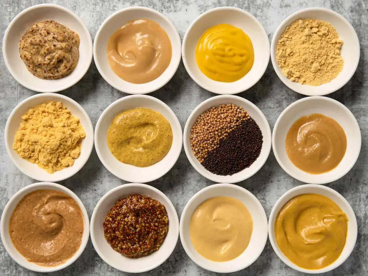 Doce recipientes blancos muestran diferentes tipos de mostaza: en grano, en polvo y en salsa, de varios tonos amarillos y marrones.