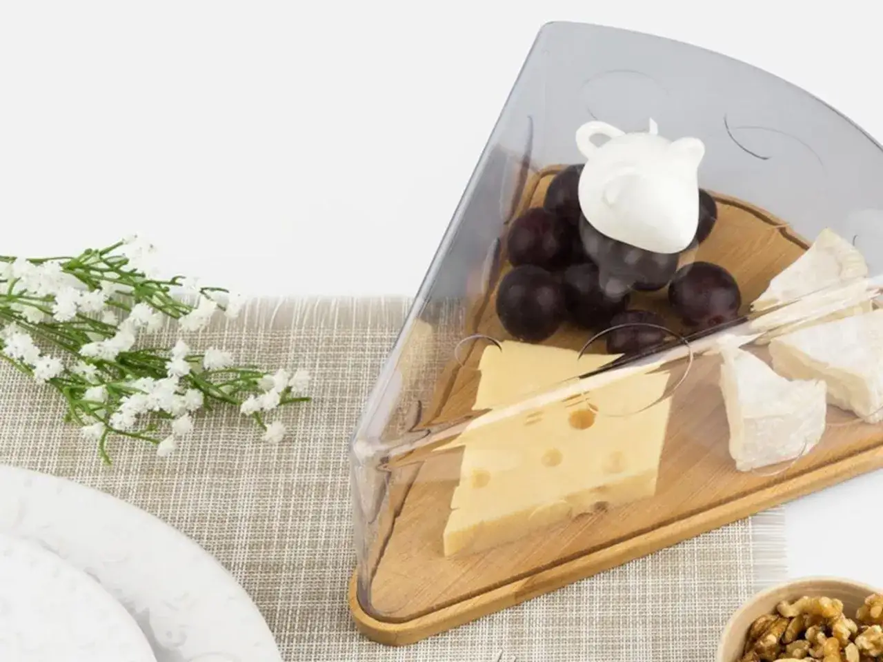 Queso, uvas y un ratón decorativo en un recipiente para conservar el queso en la nevera. Flores blancas al lado.