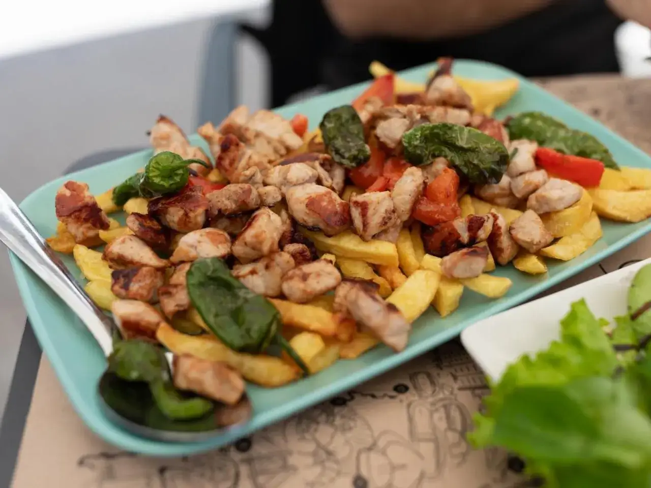 Raxo con patatas, pimientos verdes y trozos de tomate, servido en plato turquesa.