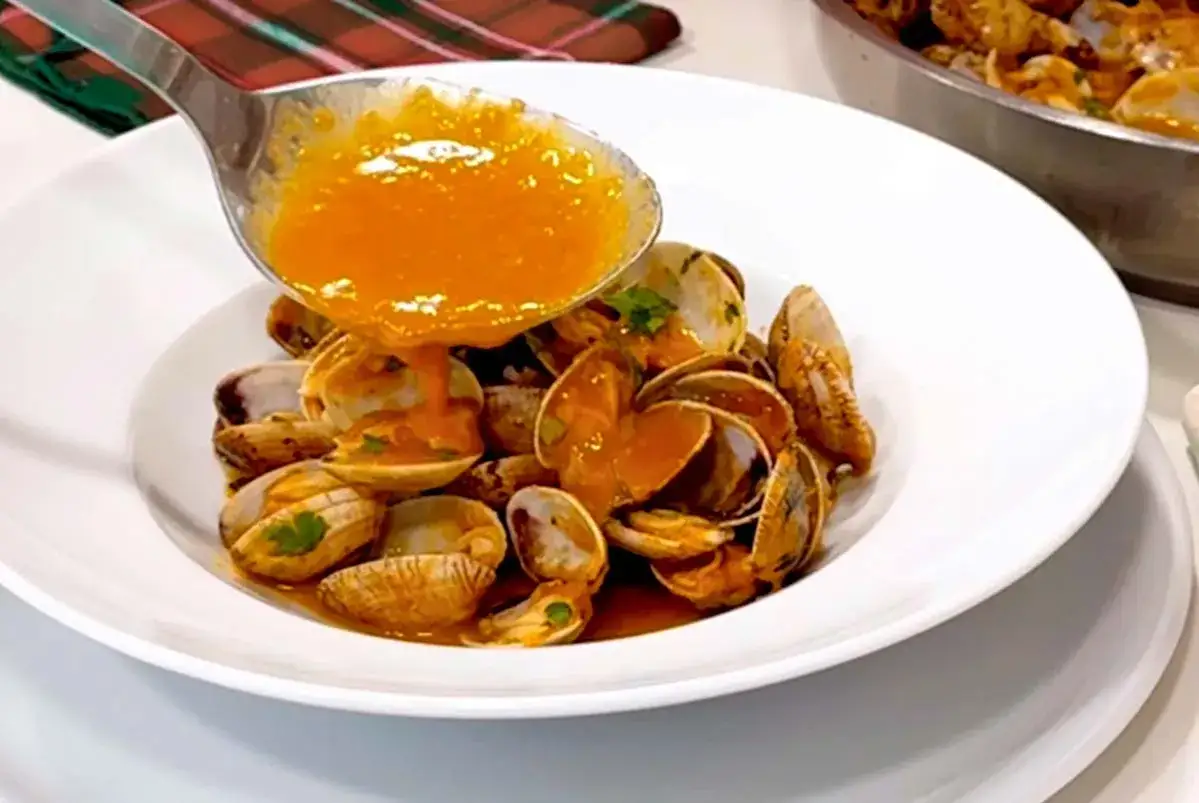 Almejas a la marinera servidas en plato blanco, con salsa naranja y perejil picado.
