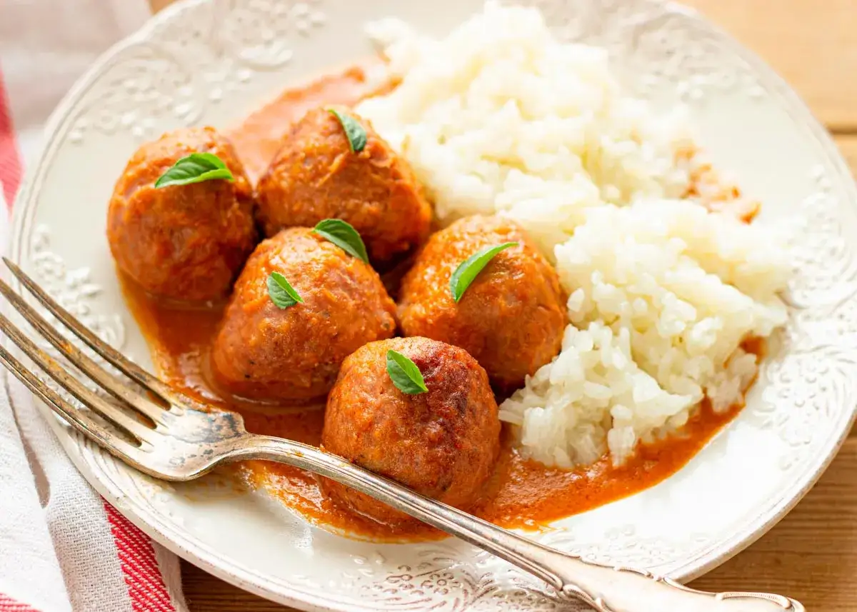Sabrosas alb&oacute;ndigas en salsa de la abuela, servidas con arroz blanco y decoradas con hojas verdes. Un plato casero que evoca recuerdos.
