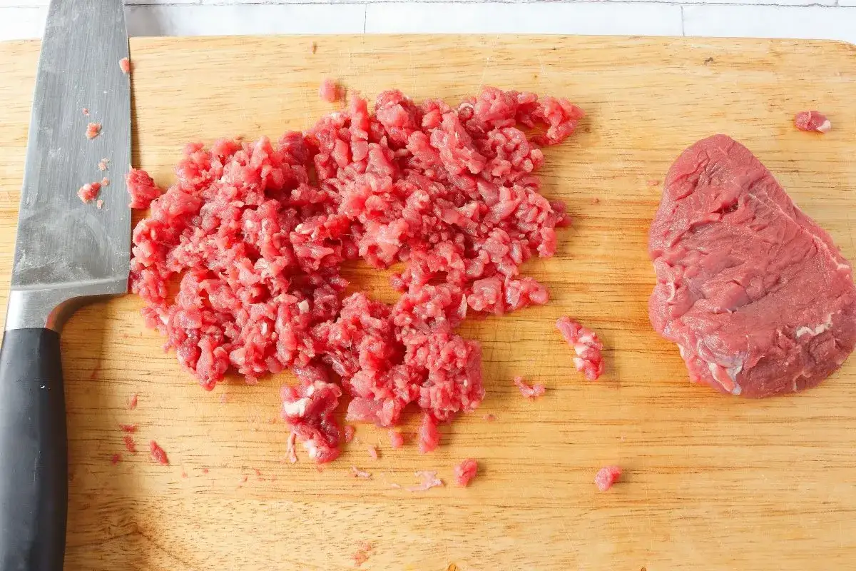 Carne picada finamente y un trozo de carne cruda en tabla de cortar, preparándose para un delicioso steak tartar.