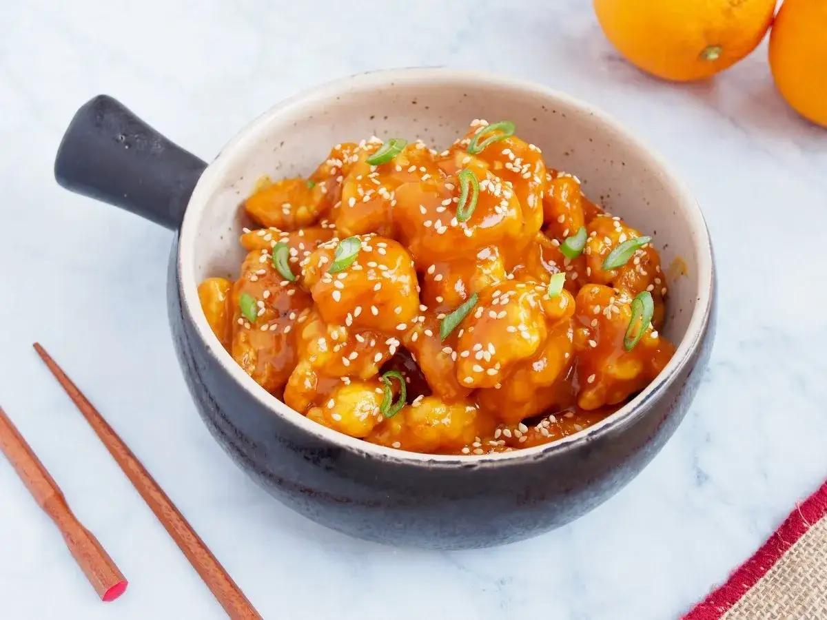 Delicioso pollo a la naranja, cubierto con semillas de sésamo y cebolleta, servido en un cuenco con palillos de madera.