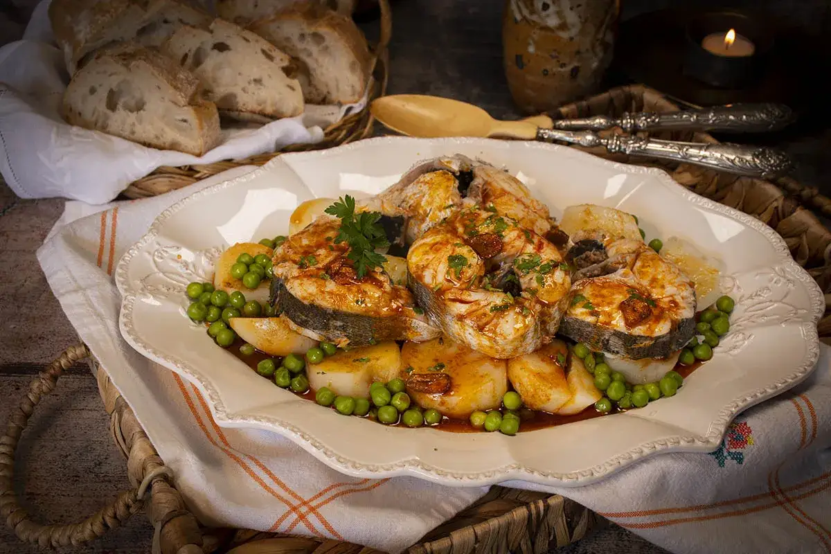 Merluza a la gallega servida en plato blanco con patatas, guisantes y perejil. Pan rústico en cesta al fondo.