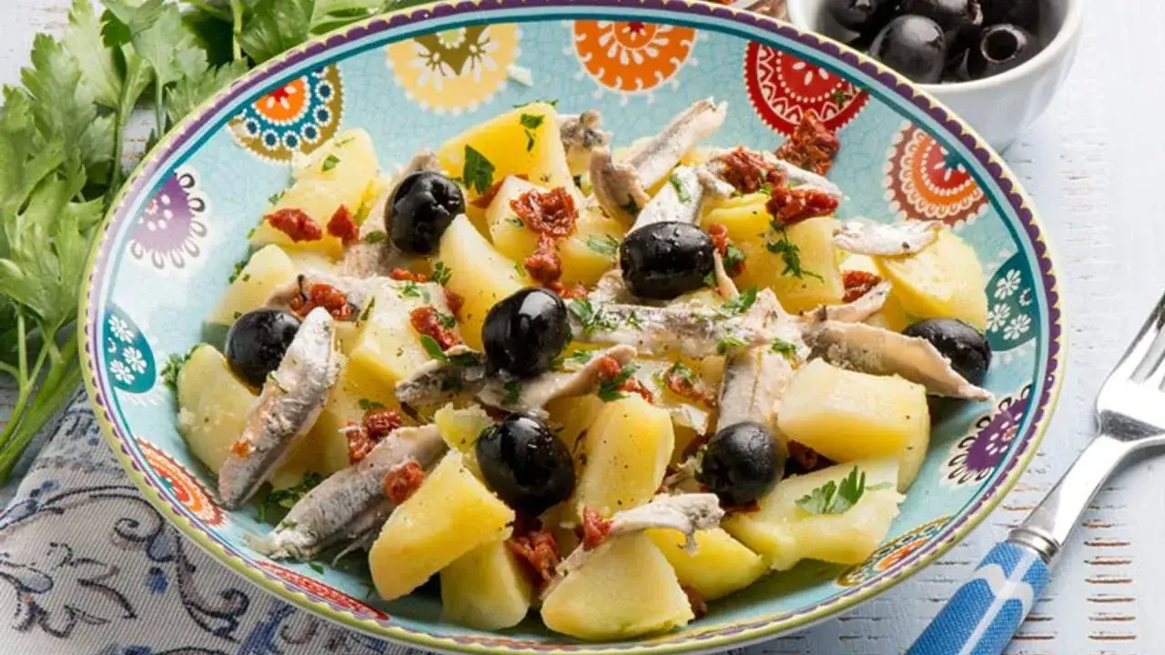 Ensalada de patatas con anchoas, aceitunas negras y tomate seco, decorada con perejil fresco.
