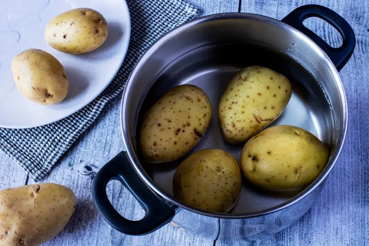 Patatas enteras en una olla con agua, listas para cocer. Otras patatas reposan en un plato y sobre la mesa.