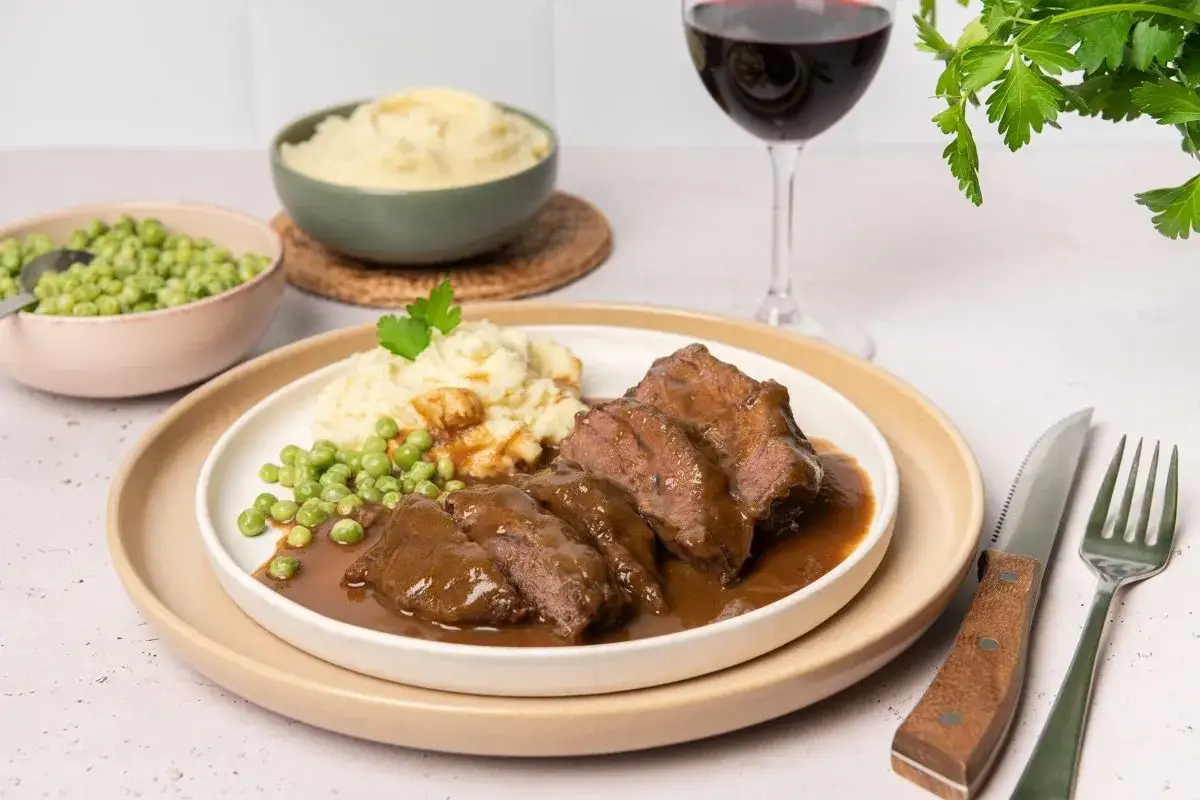 Carrilleras de ternera estofadas en salsa, servidas con pur&eacute; de patatas y guisantes. Un plato reconfortante acompa&ntilde;ado de vino tinto.