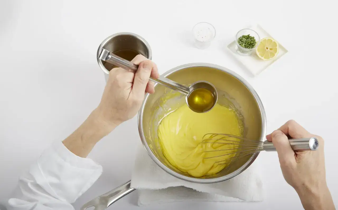 Manos expertas logran la emulsión perfecta en cocina, añadiendo aceite a una mezcla amarilla con un batidor.