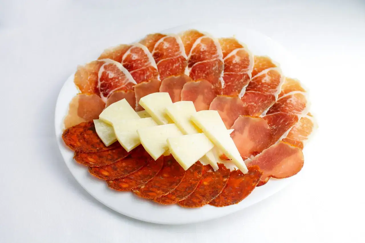 Una tabla de quesos como hacerla: jamón serrano, chorizo y queso en cuña, dispuestos en un plato blanco.