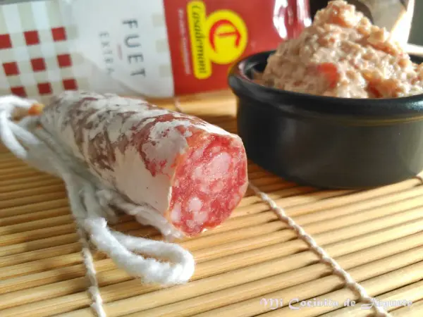 Un trozo de fuet extra con su interior rosado y veteado, junto a un cuenco de paté.